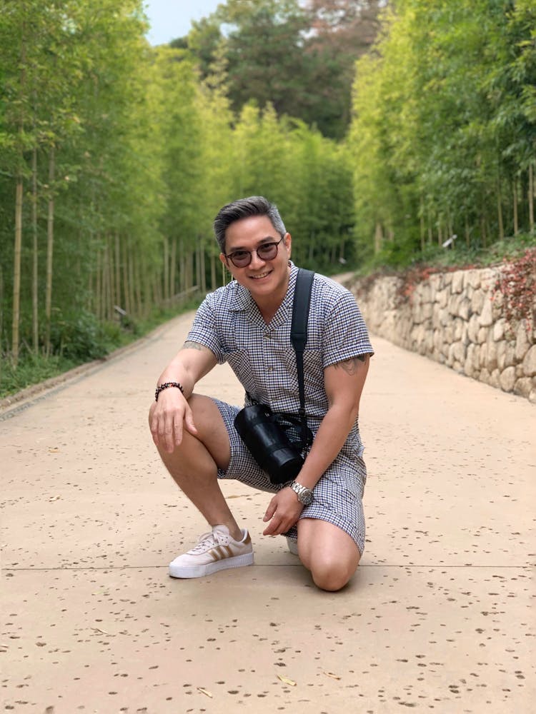 A Smiling Man Wearing Onesie And Black Sling Bag While Sitting On The Street Between Green Trees