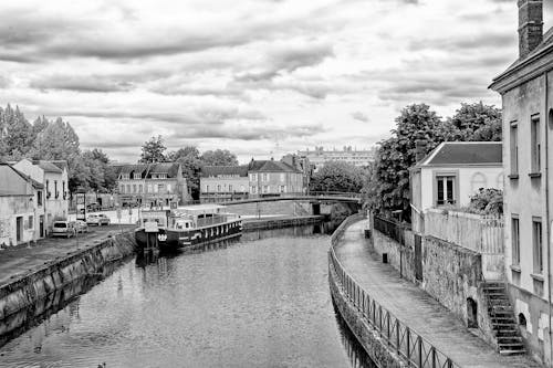 A Grayscale Photo of a River Between Houses Under the Cloudy Sky