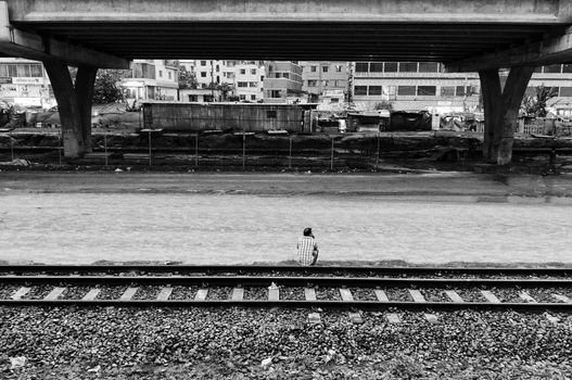 A black and white image of a man sitting on train tracks under an urban bridge, evoking solitude.
