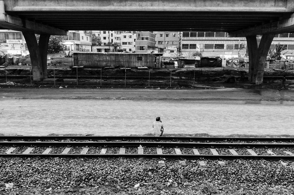 A black and white image of a man sitting on train tracks under an urban bridge, evoking solitude.