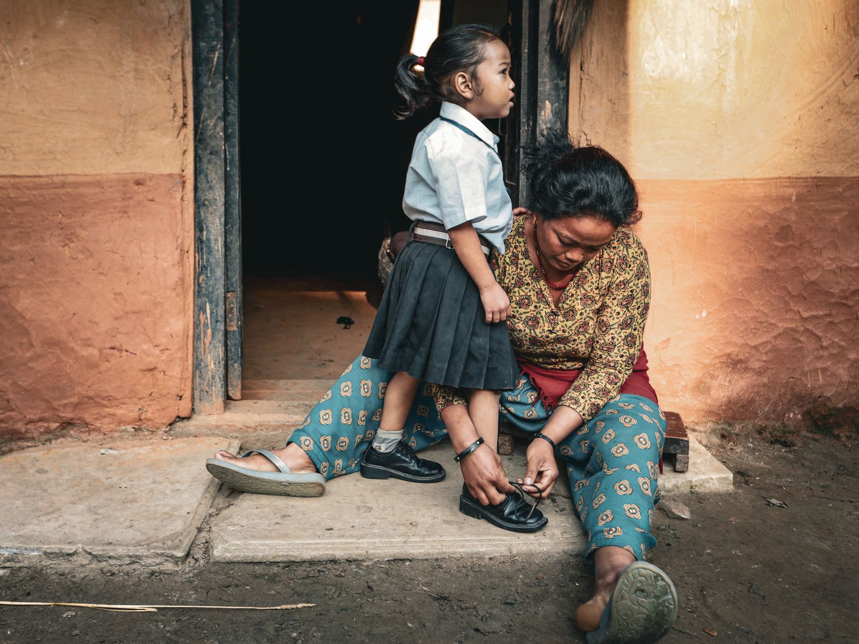 Mother tying daughter's shoes at doorstep, ready for school day.