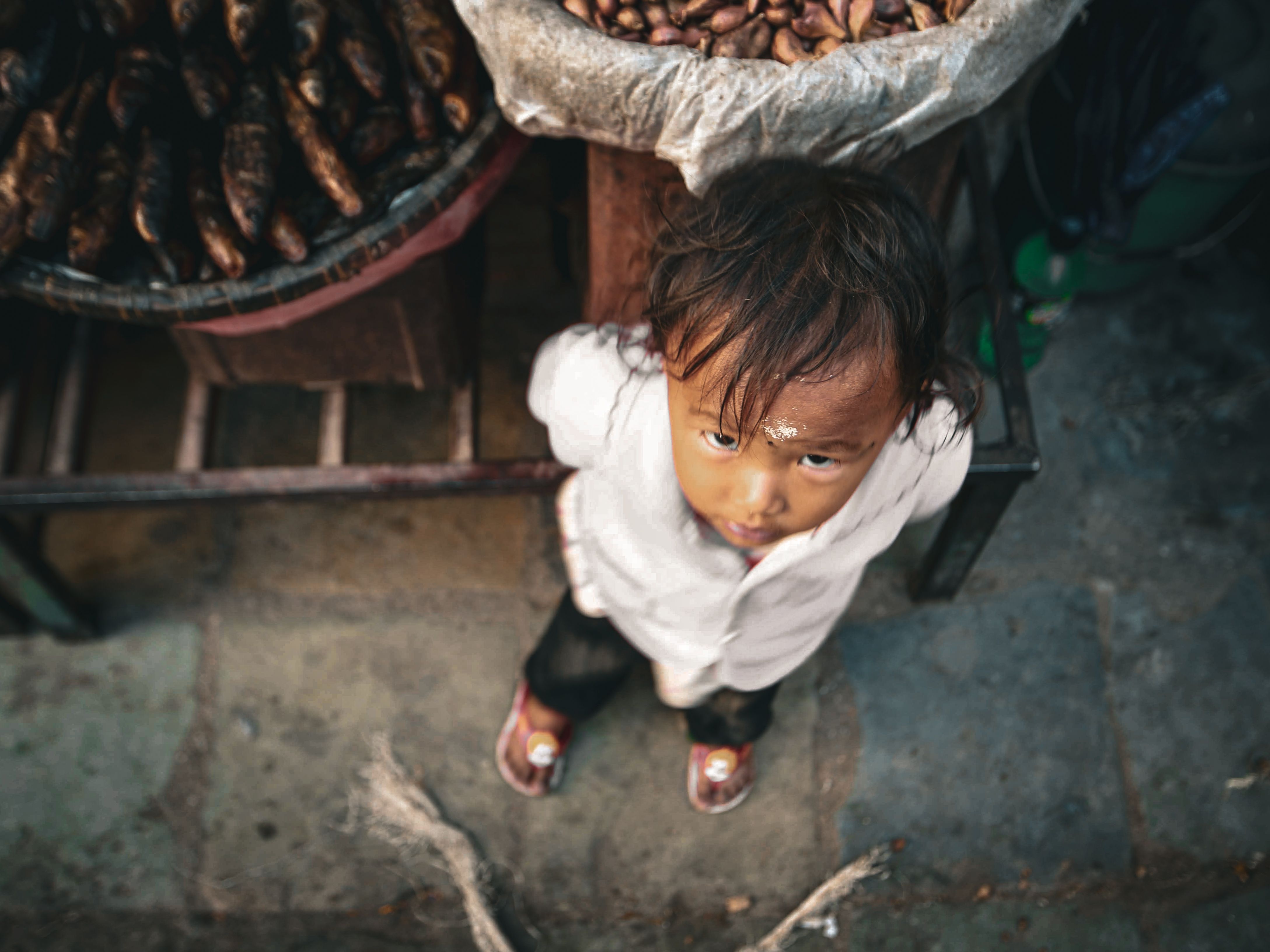 A curious child looks up at the camera while standing near a basket of vegetables in an outdoor market setting.