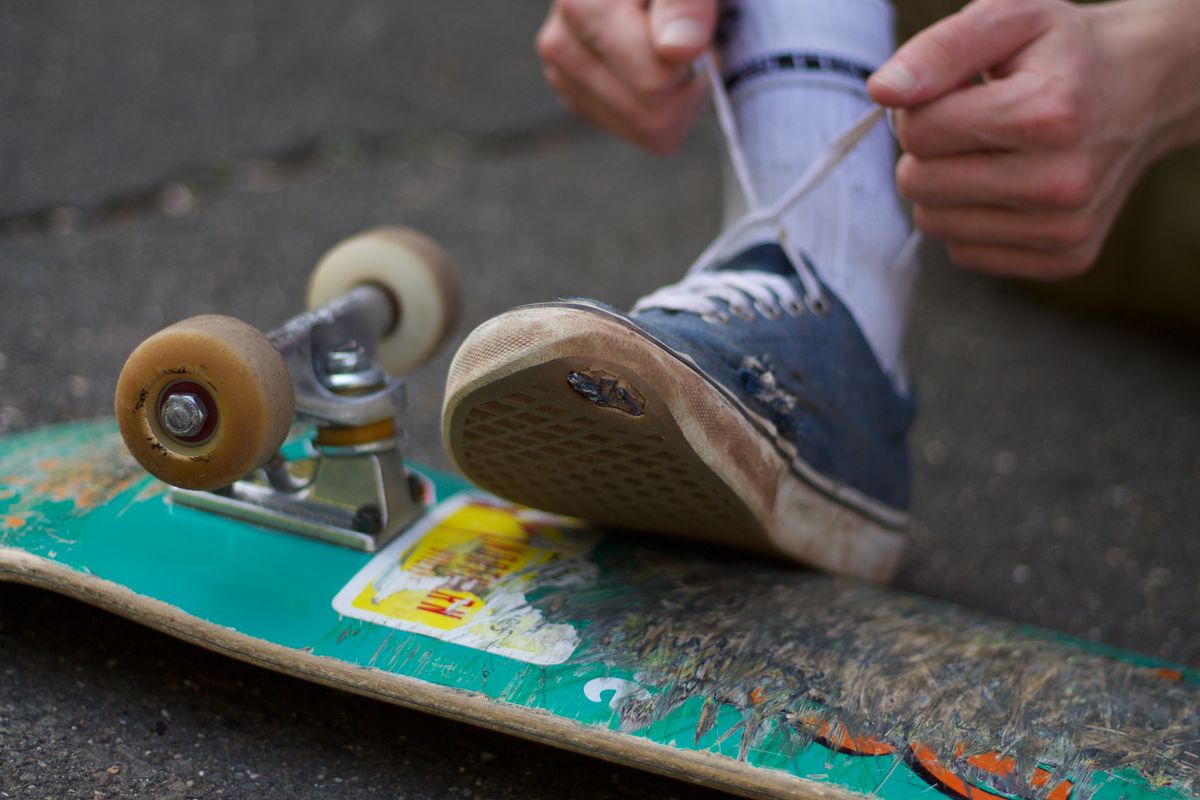 A worn skate shoe with a scuffed, flattened sole resting on a skateboard while its owner ties the laces.