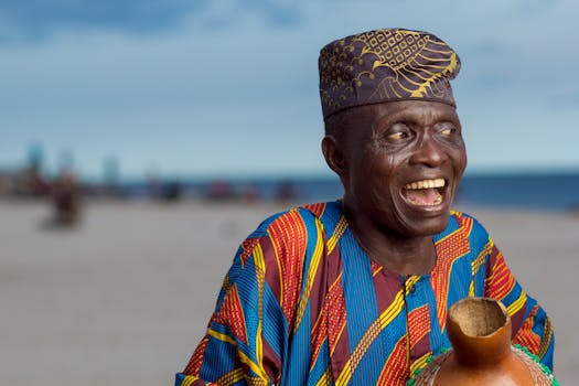 Elderly man joyfully smiling wearing Yoruba attire and cap on Lagos beach.