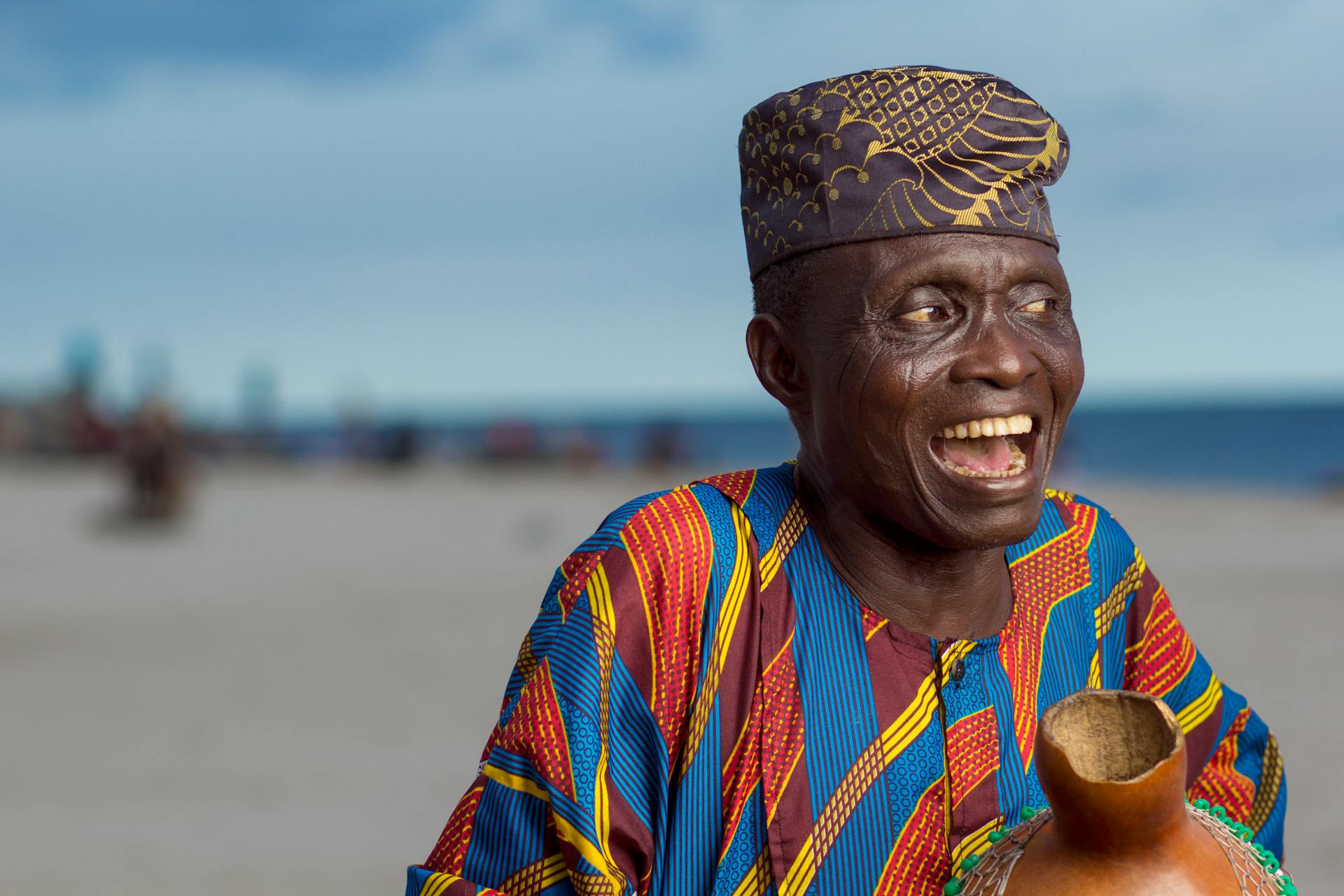 Elderly man joyfully smiling wearing Yoruba attire and cap on Lagos beach.