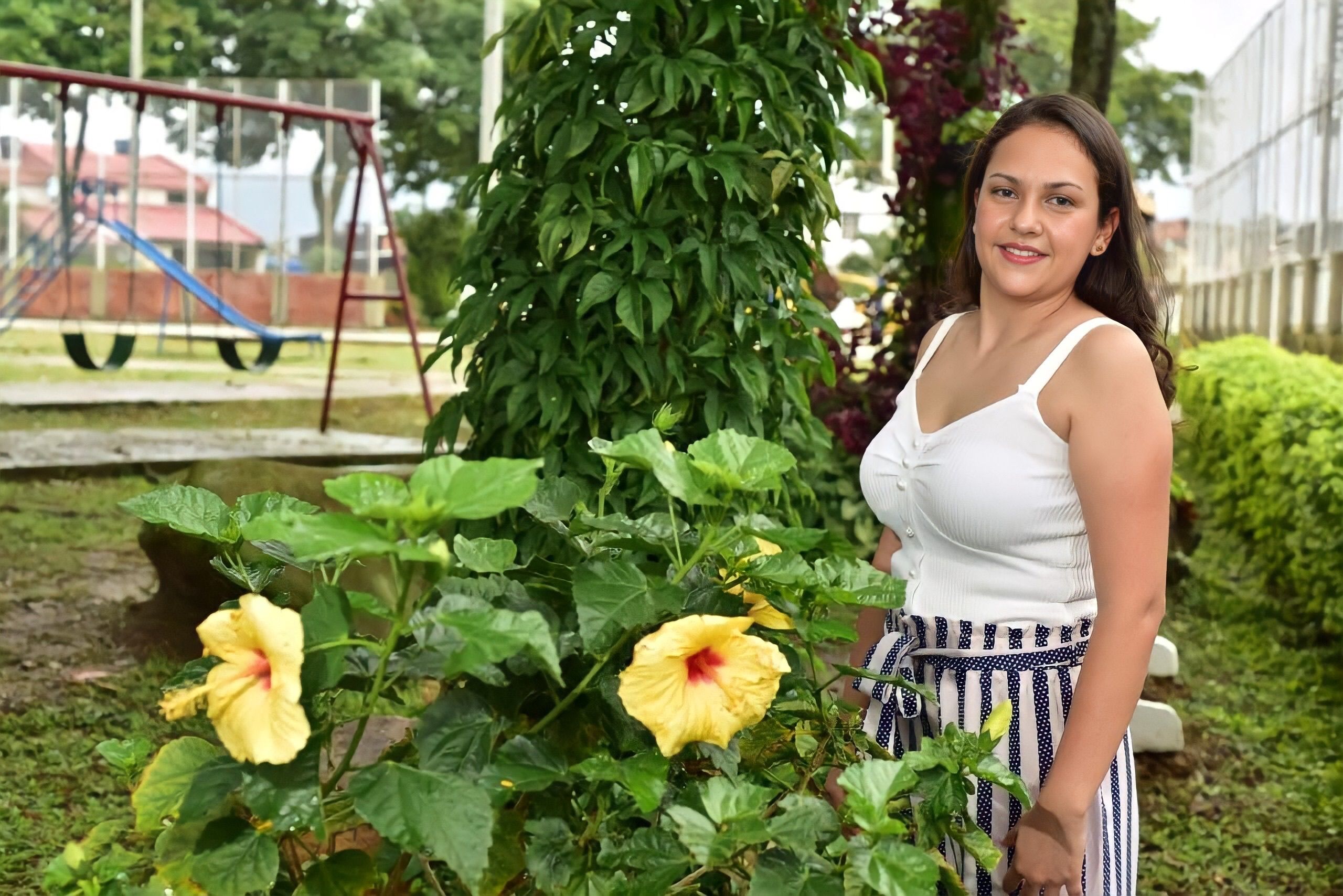 Woman standing in a vibrant garden with yellow hibiscus flowers and a playground in the background.
