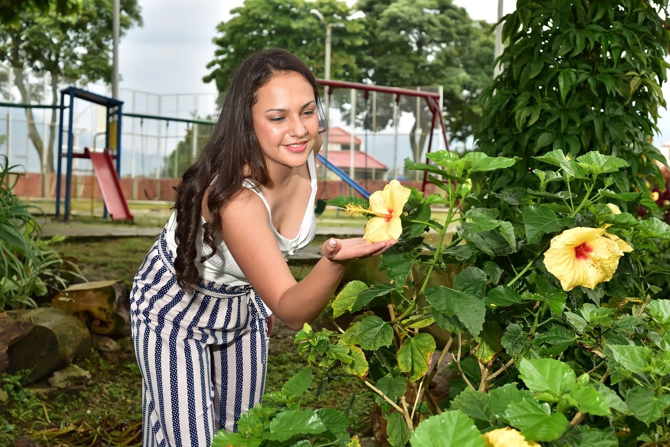 A woman in a garden smiling while holding a yellow hibiscus flower in a park setting.