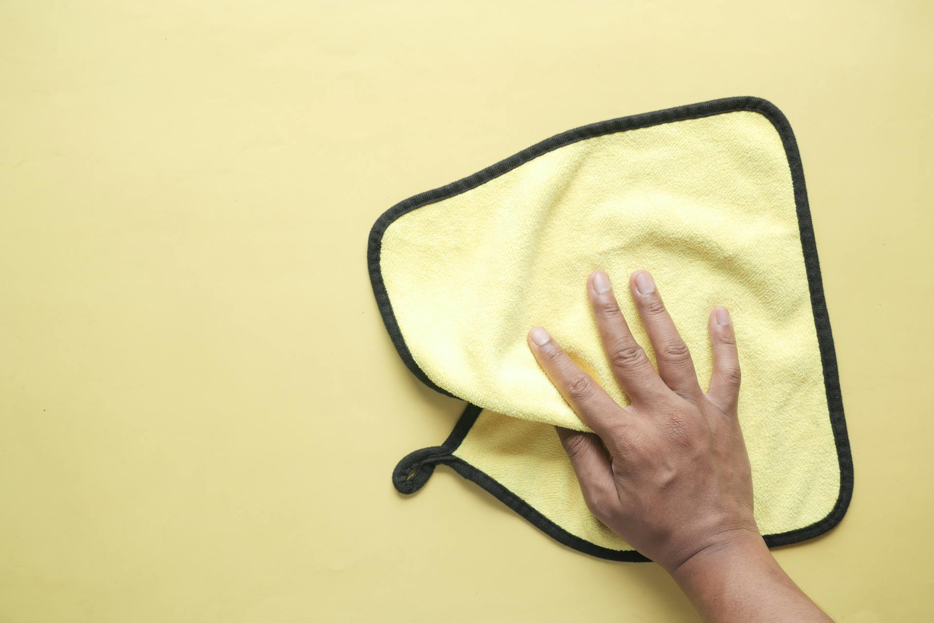 A hand using a microfiber cloth to wipe a yellow background, demonstrating cleaning and hygiene concepts.