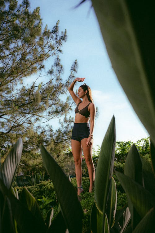 Posed Photo of a Woman in a Bra and Shorts Raising her Hand and Standing among Green Plants and Trees