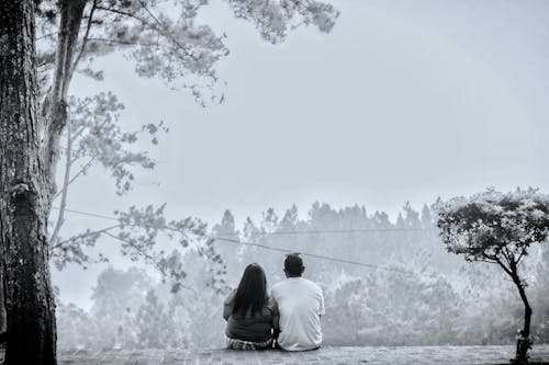 Grayscale Photo of Couple Sitting Near the Trees 