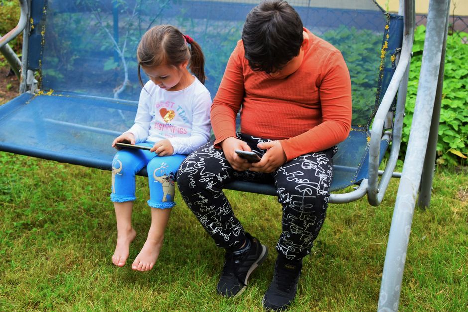 Two children using mobile phones while sitting on a swing outdoors in a garden.