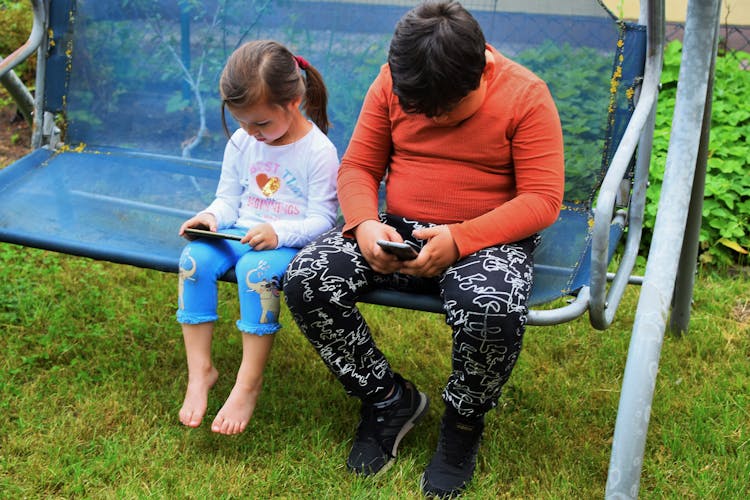 Children Sitting On Swing In Garden Using Cellphones