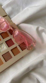 A trendy eyeshadow palette paired with pink sunglasses on a soft white fabric, perfect for fashion and beauty themes.