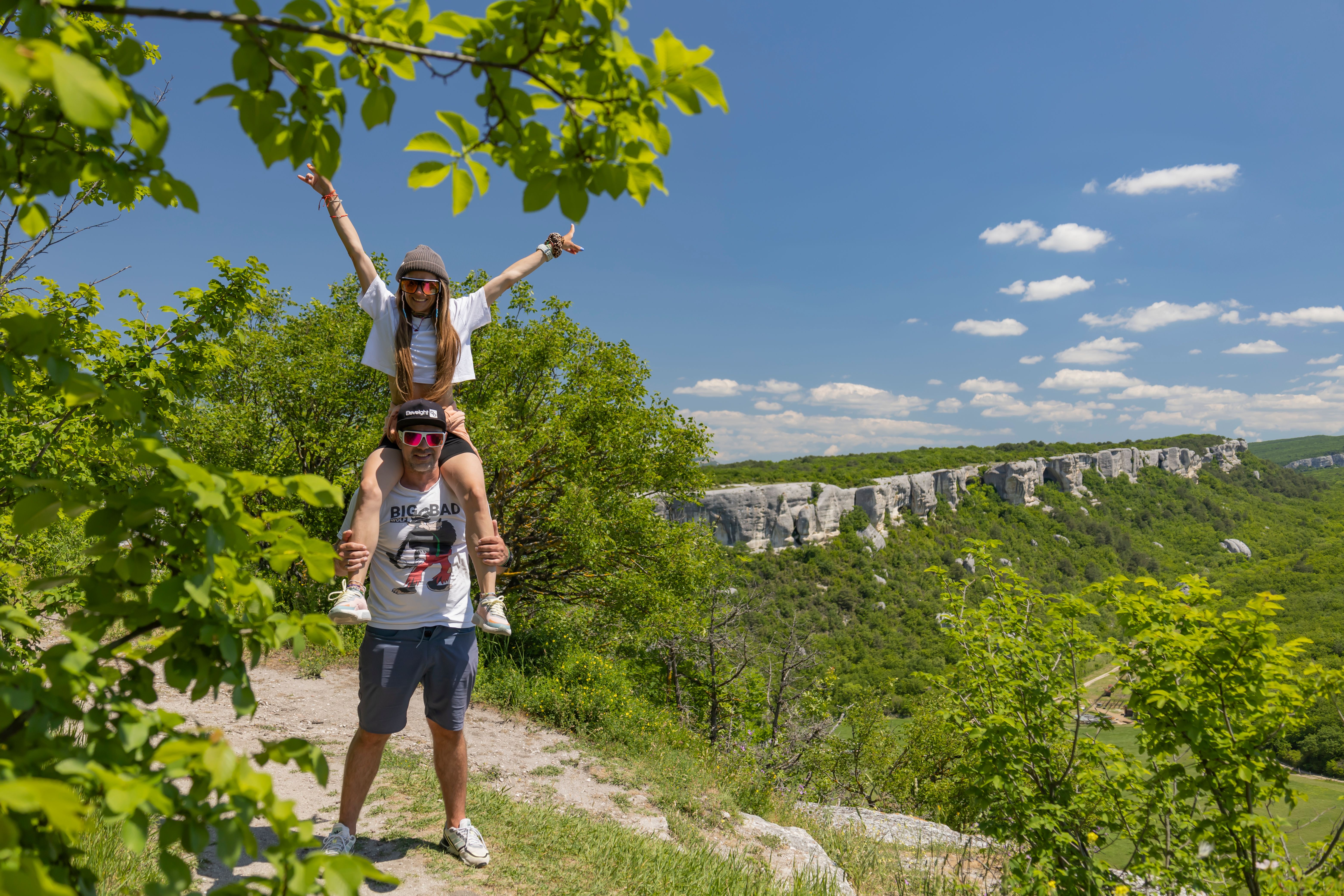 A couple enjoys a playful piggyback ride on a scenic countryside hike under a sunny sky.