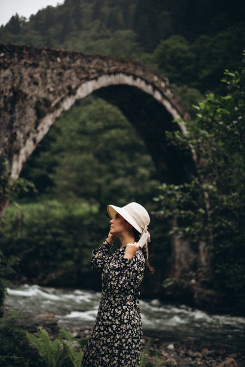 Woman in Straw Hat and Floral Pattern Dress Posing in front of an Old Arched Bridge