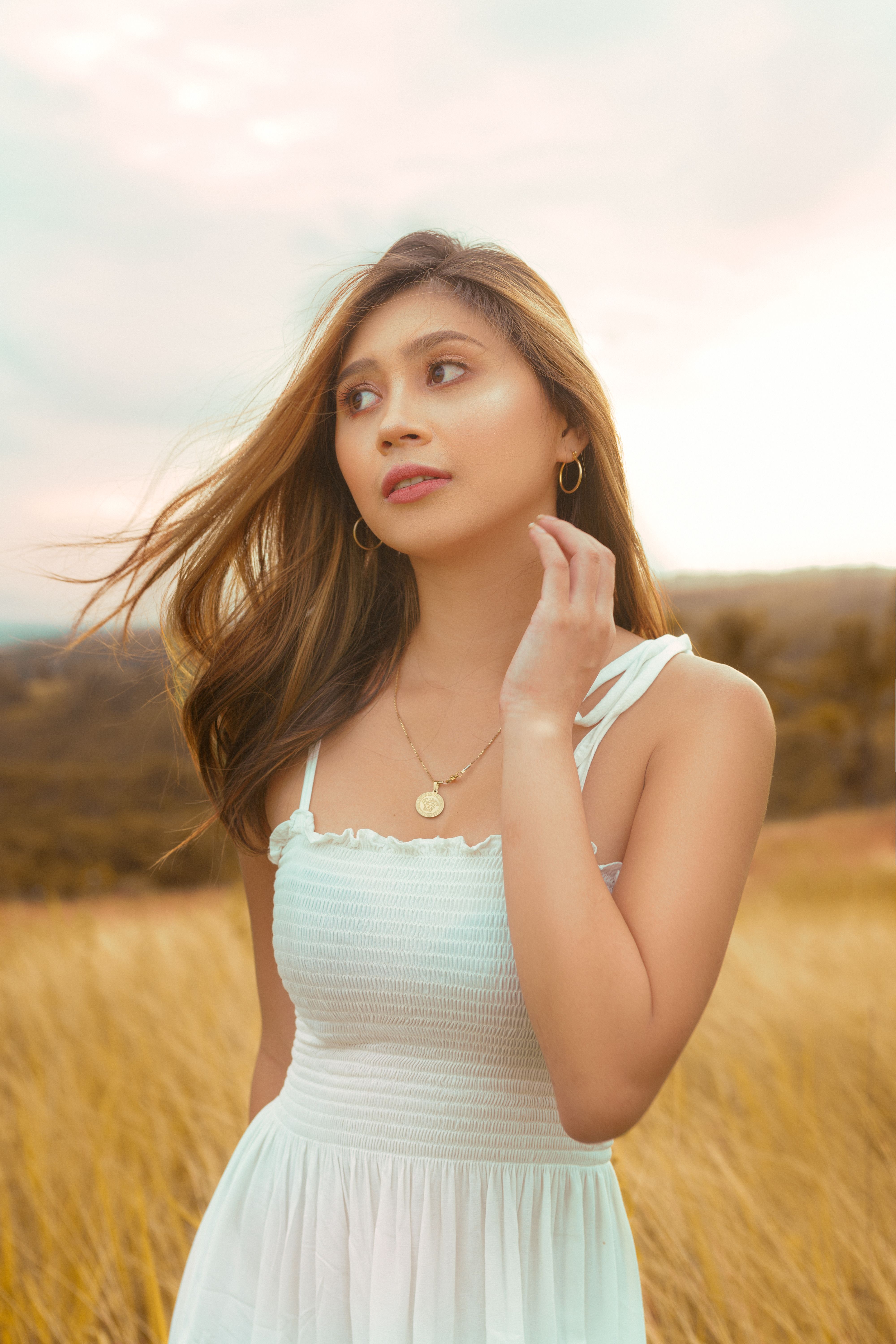 A woman in a white dress stands thoughtfully in a golden field during sunset.