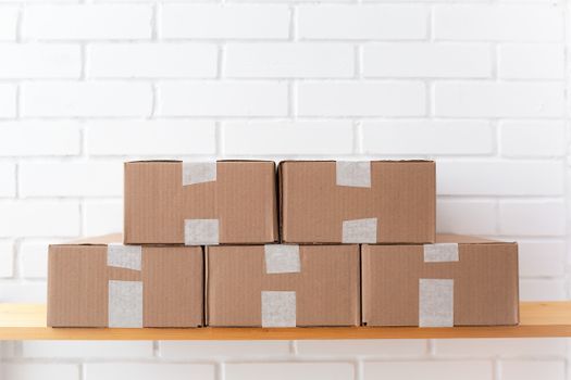 A neat stack of cardboard boxes taped and placed on a wooden shelf against a white brick wall.