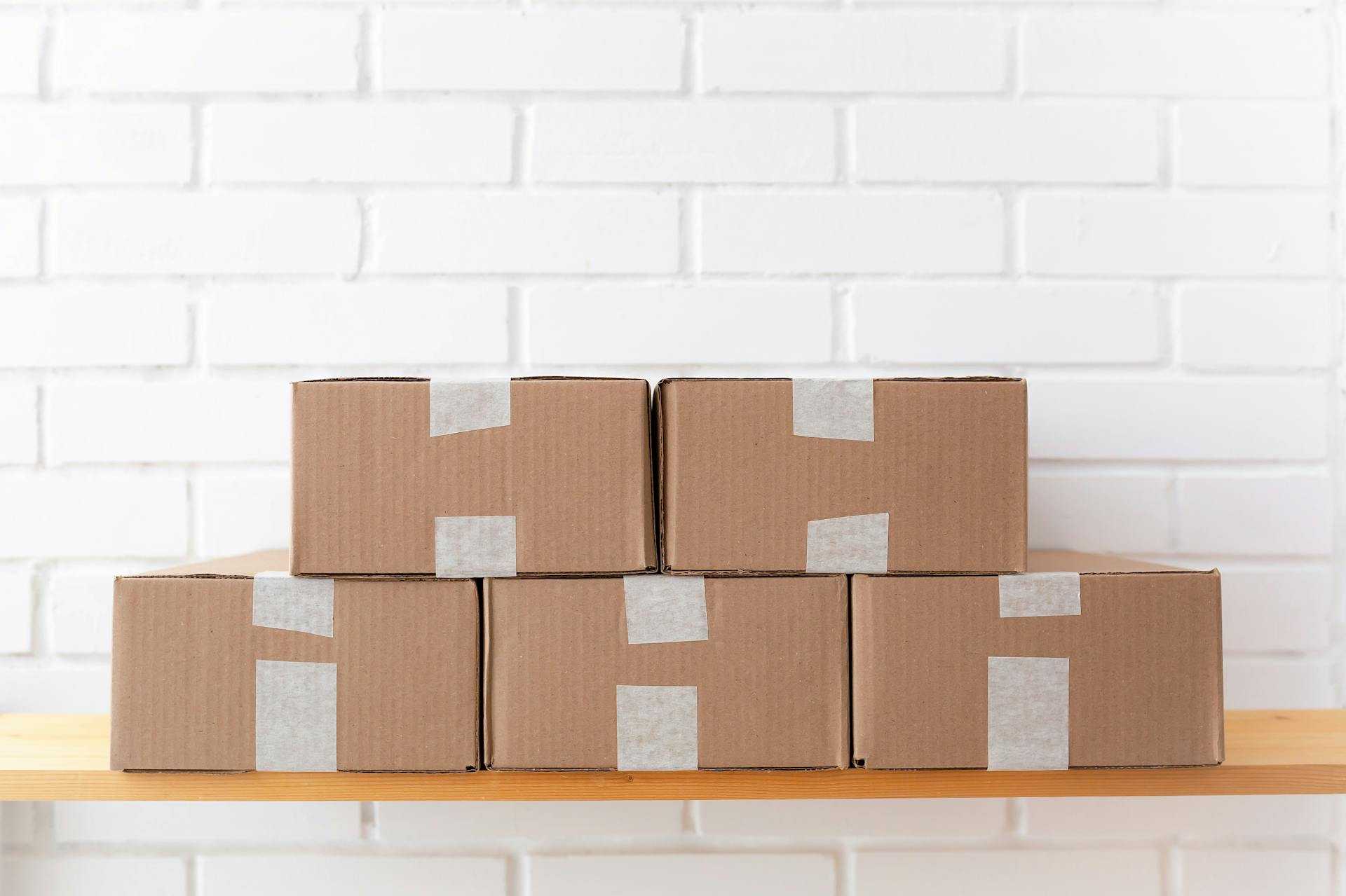 A neat stack of cardboard boxes taped and placed on a wooden shelf against a white brick wall.