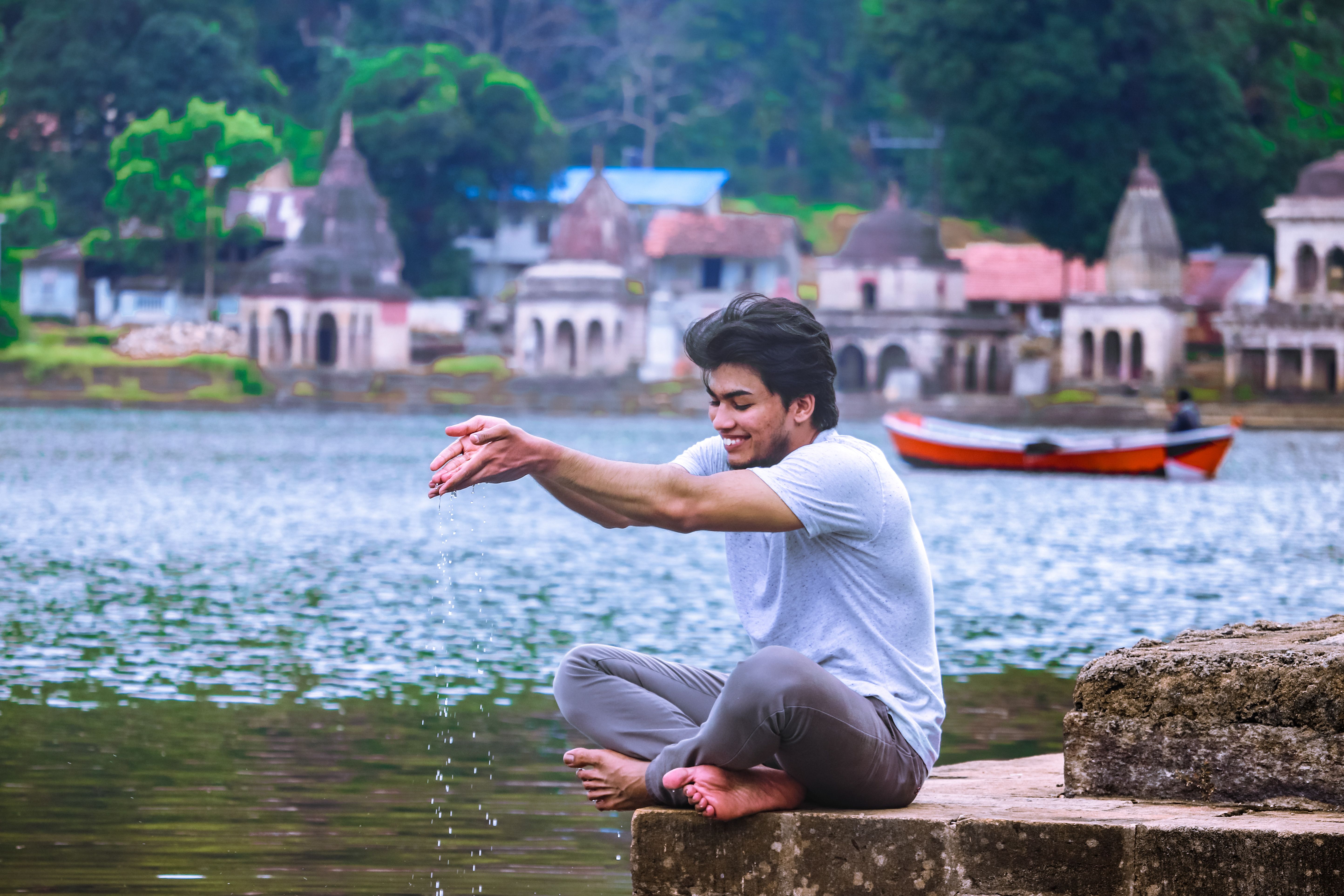 Young man sitting by a riverside in India, playing with water and smiling, capturing a serene moment.