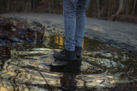 A person in jeans and boots standing on a puddle causing ripples with reflection.