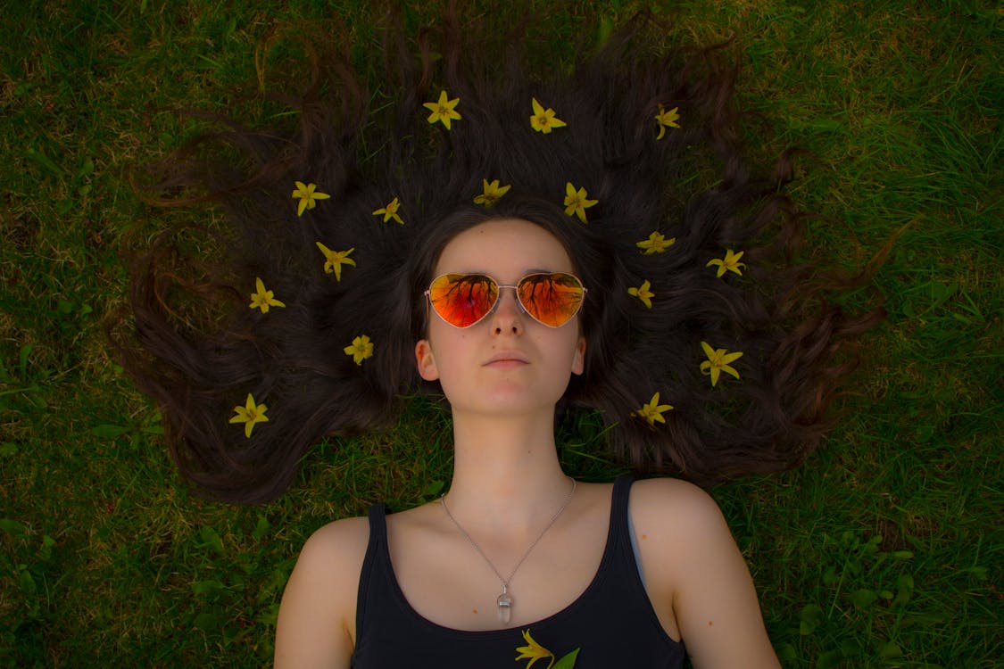 Photo of Lying Woman on Green Grass Wearing Red Lens Heart Sunglasses With Yellow Petal Flowers on Hair