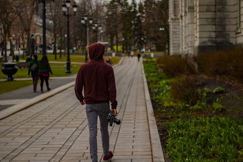 コンクリートの建物の近くでデジタル一眼レフカメラを保持しているコンクリートの経路を歩くパーカーを着た男