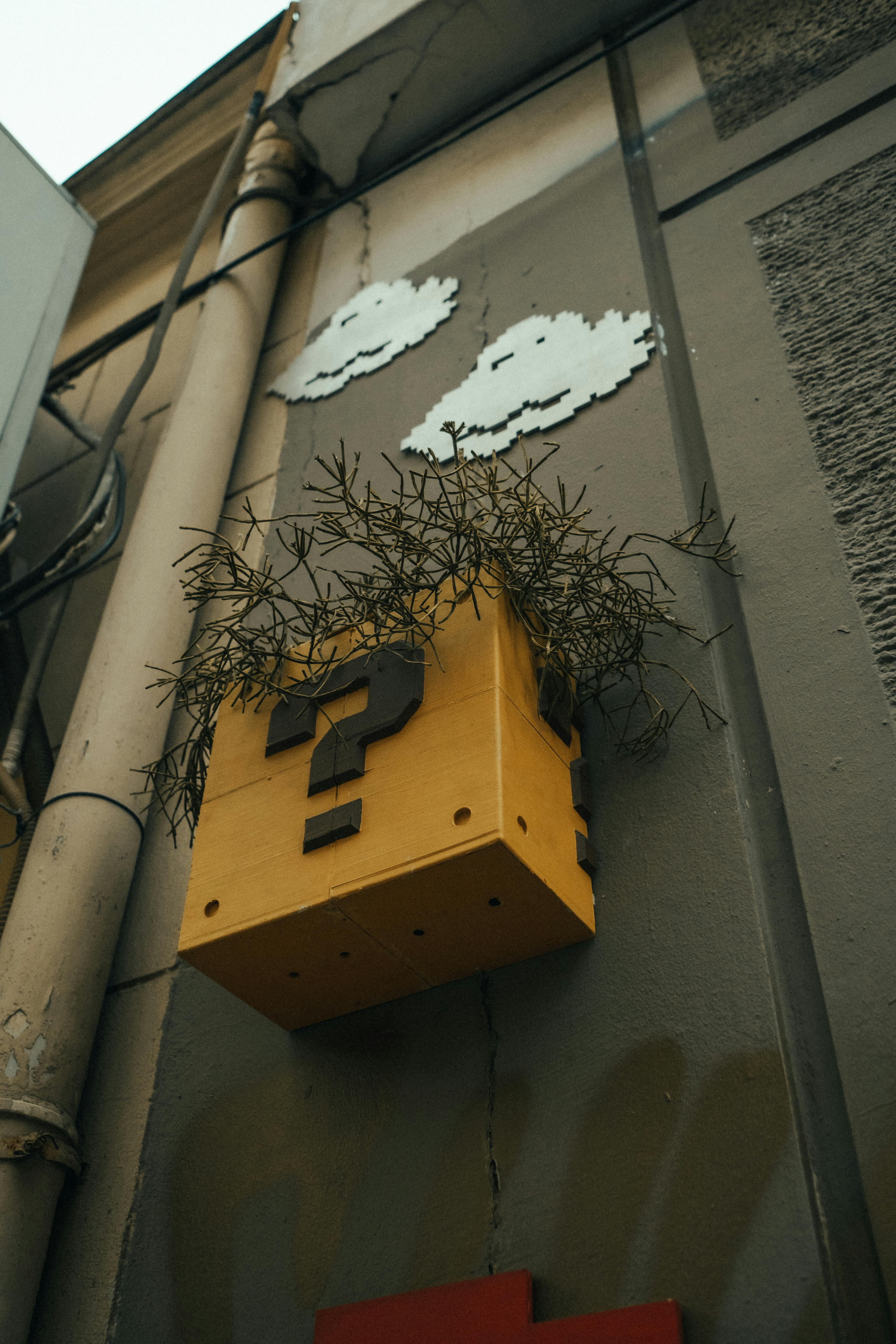 Street art featuring a yellow question block with plants on a building wall under metal clouds.