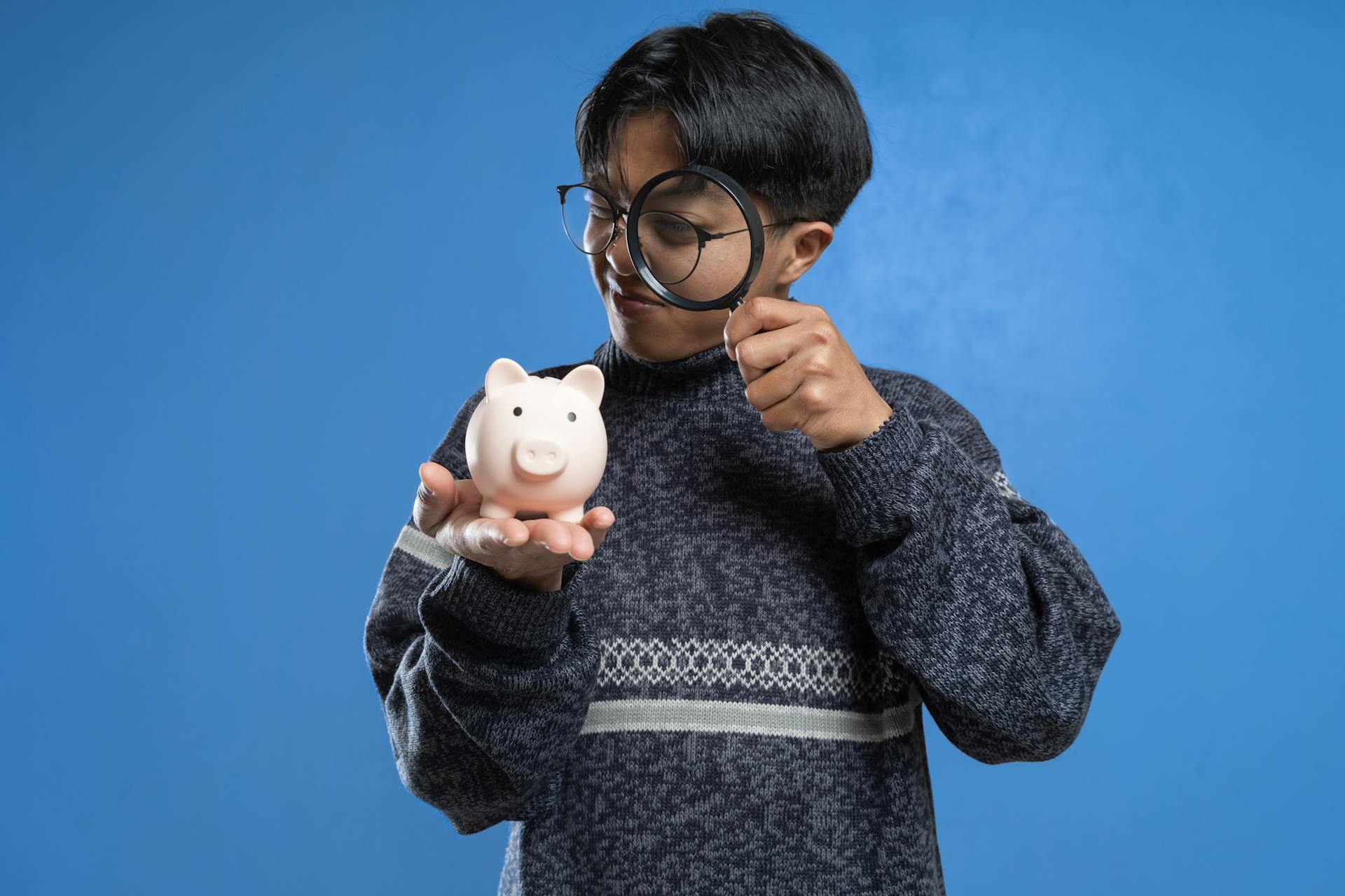 A person closely examines a piggy bank with a magnifying glass, symbolizing financial scrutiny.