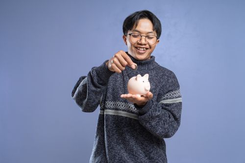 Free Smiling man holding a piggy bank and inserting a coin, representing savings and financial planning. Stock Photo