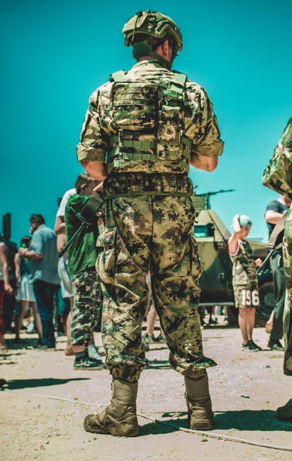 A soldier in military camouflage standing outdoors, surrounded by people and vehicles.