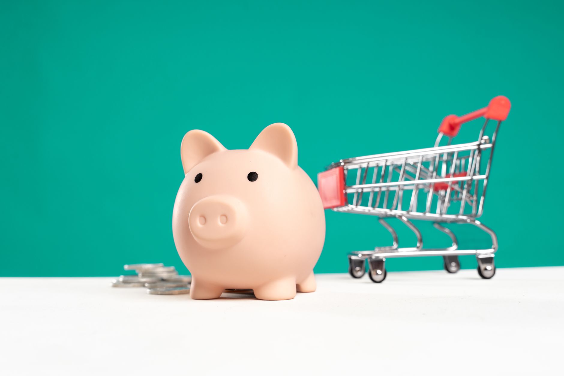 Piggy bank with shopping cart and coins on a white and teal background, symbolizing saving and spending.