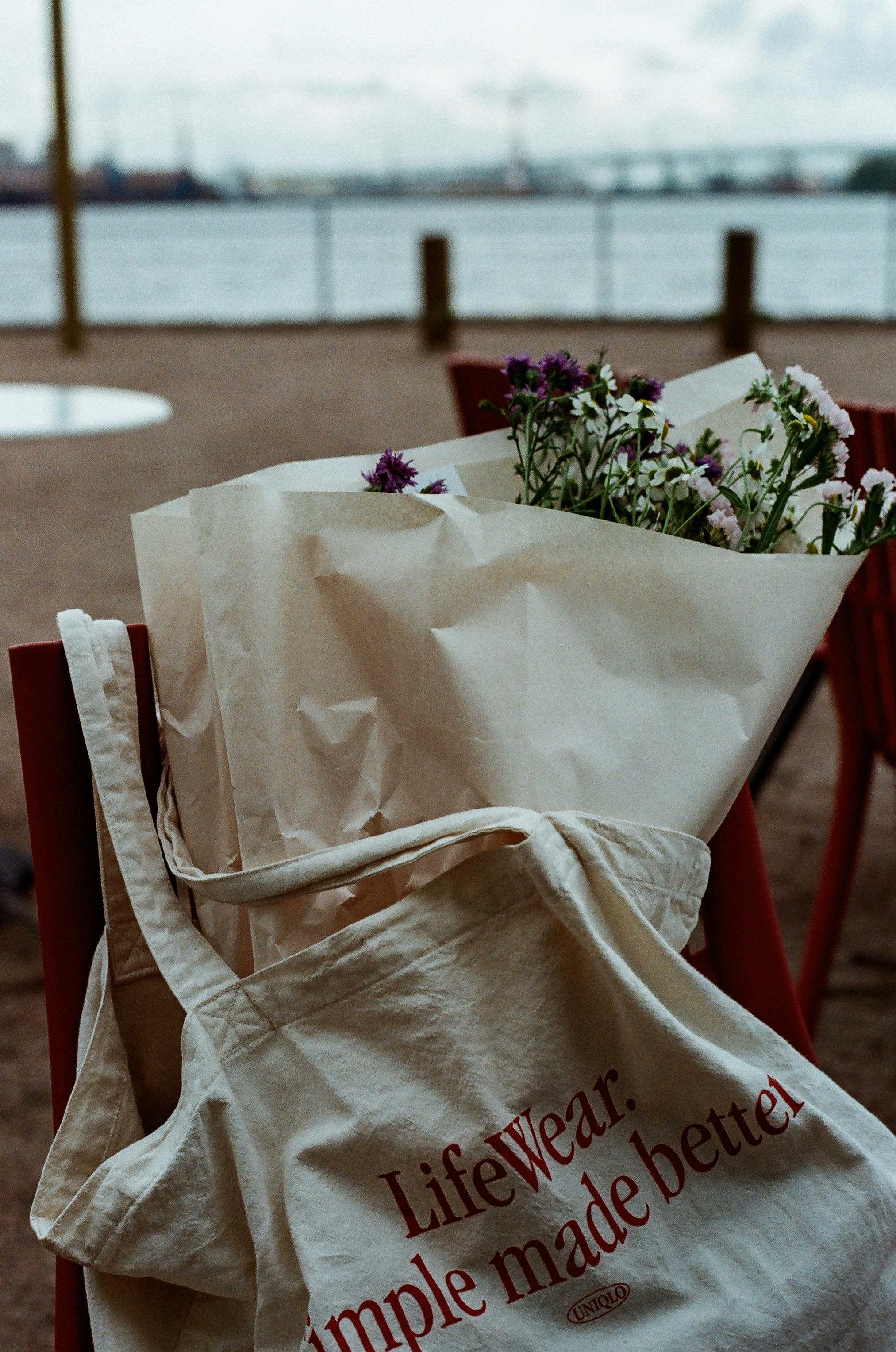 A minimalist outdoor scene with a tote bag and a bouquet of wildflowers on a chair by the water.