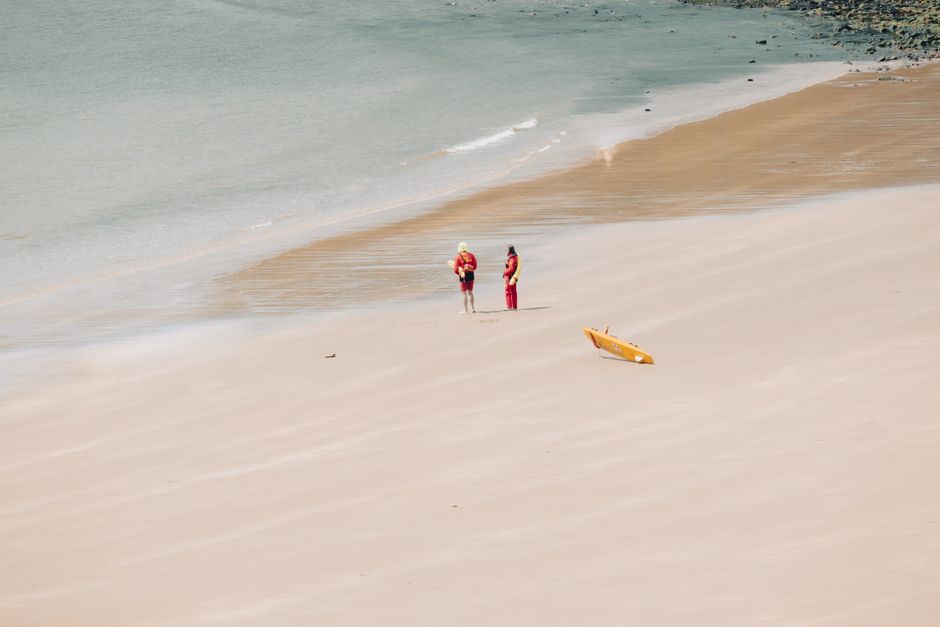 Two lifeguards on a sandy beach near the ocean with a surfboard nearby.