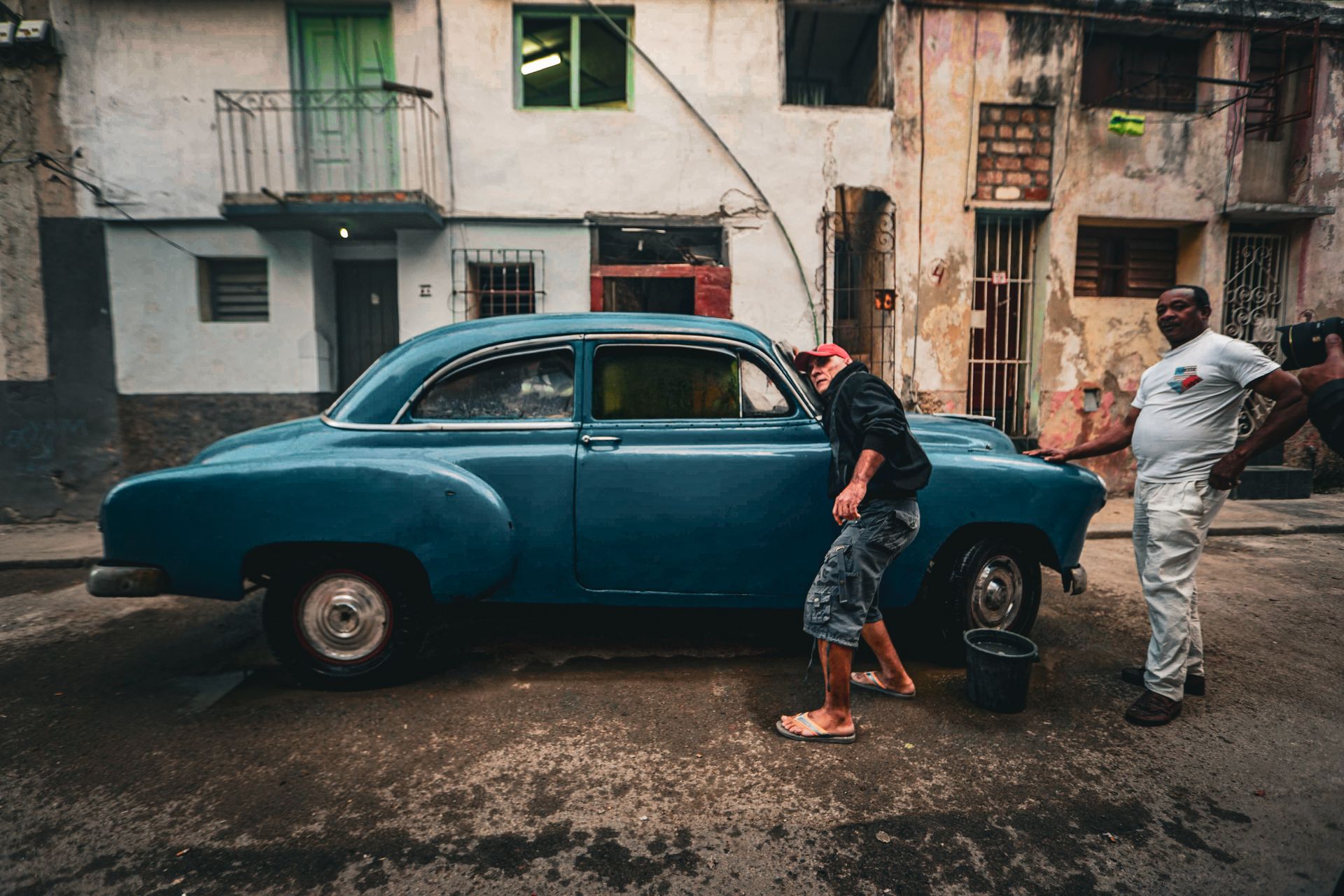 Two men cleaning a vintage car on a street lined with rustic buildings.
