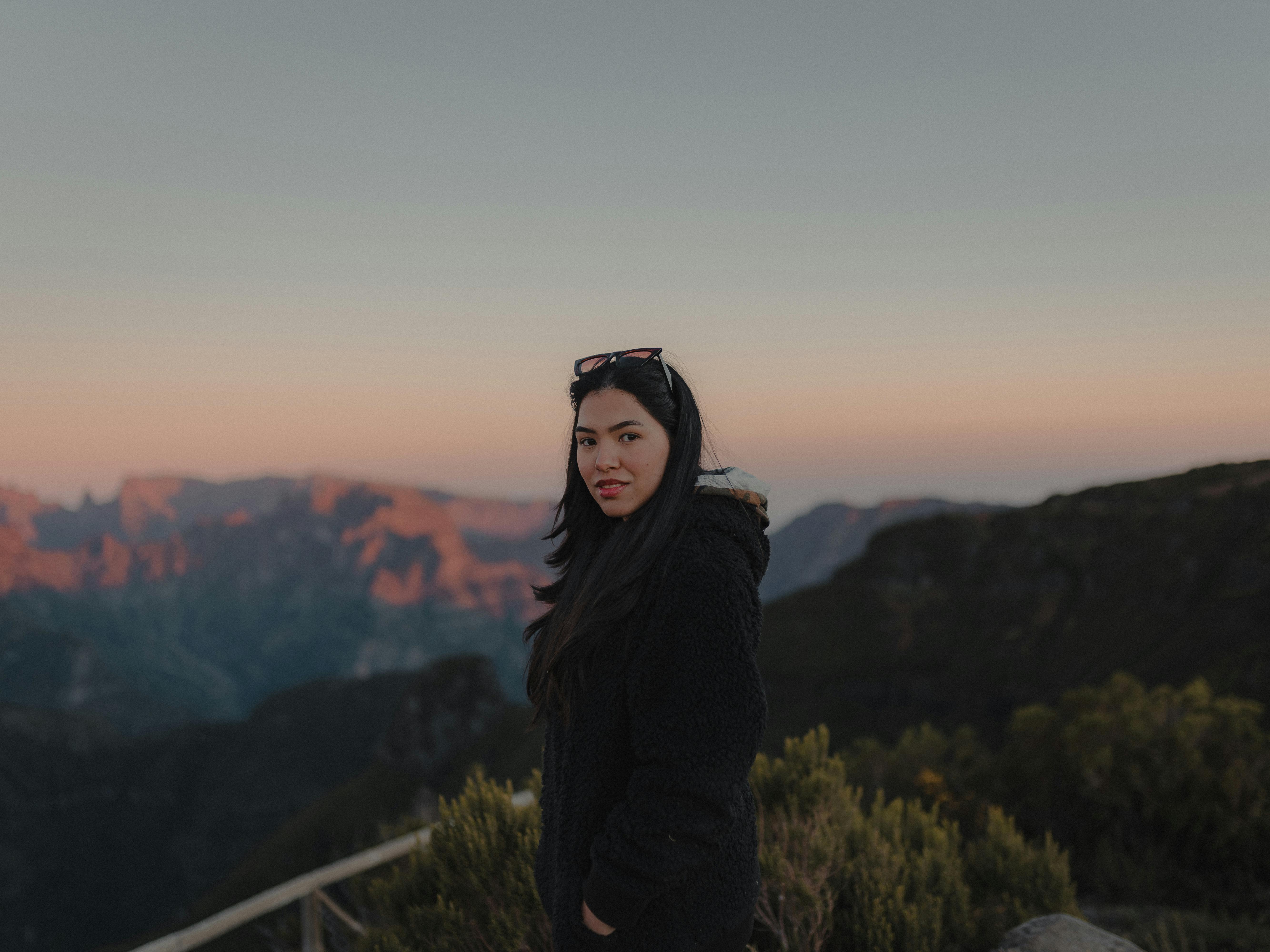 A woman in a coat stands in the mountains at sunset, capturing serene beauty.