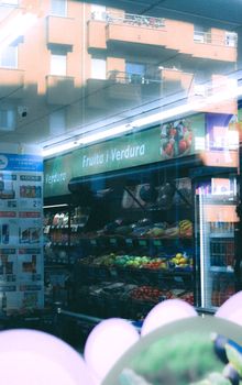 A vibrant produce aisle in a grocery store viewed through a reflective window, showcasing fresh fruits and vegetables.