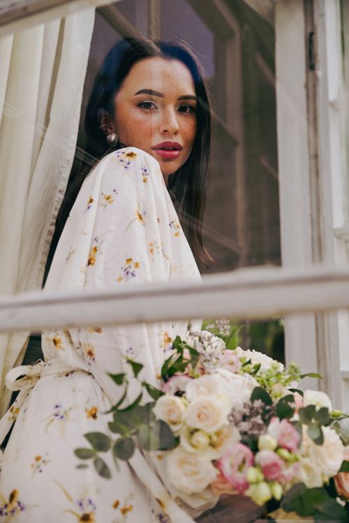 Free Elegant woman in floral dress holding a bouquet, looking through a window. Stock Photo