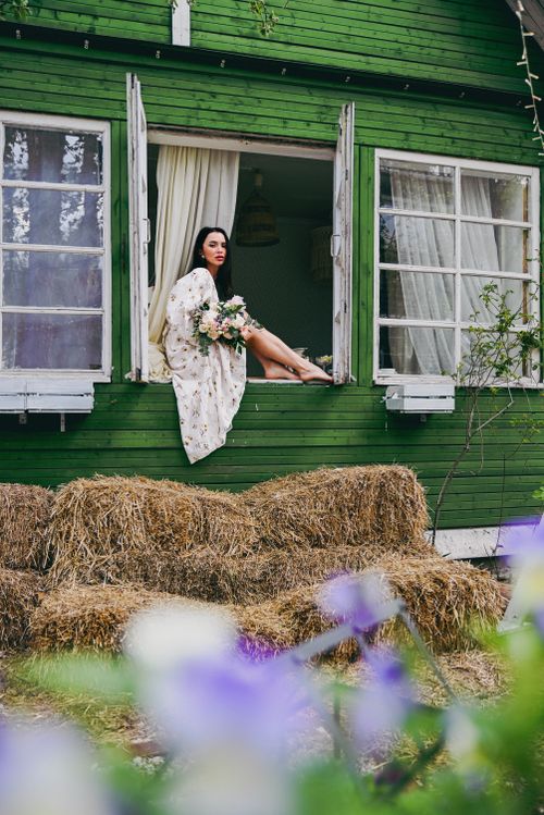 Free Woman in floral dress sitting in window of a green house surrounded by hay bales and flowers. Stock Photo
