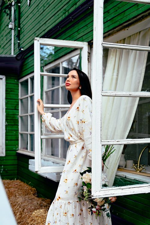 Free Woman in a floral dress holding a bouquet, looking contemplatively outside a green wooden house. Stock Photo