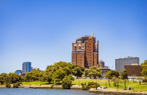 Free A tall historic brick building overlooking a park and waterfront under a clear blue sky. Stock Photo