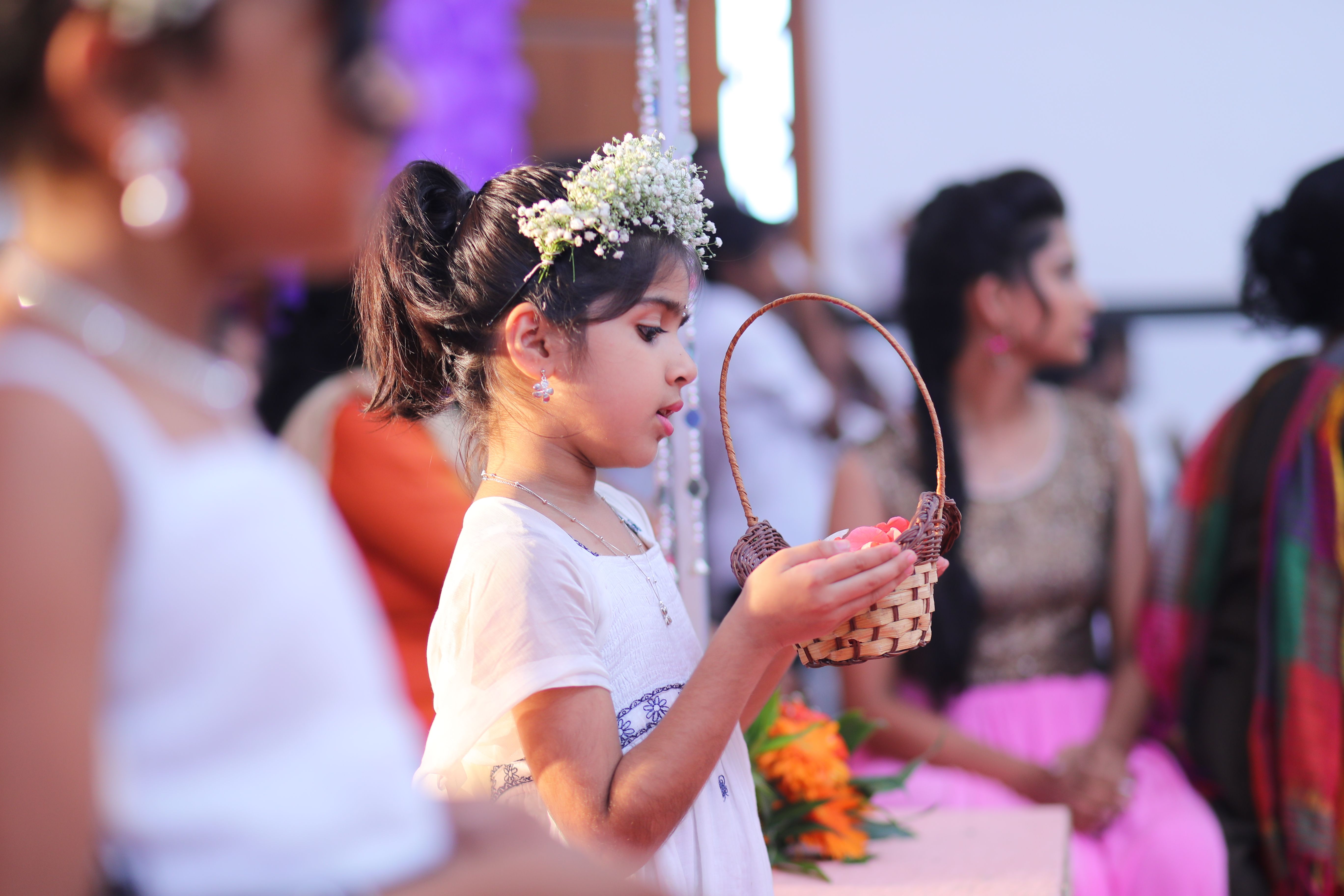 A young flower girl in a white dress holds a basket at a ceremony, surrounded by vibrant colors.