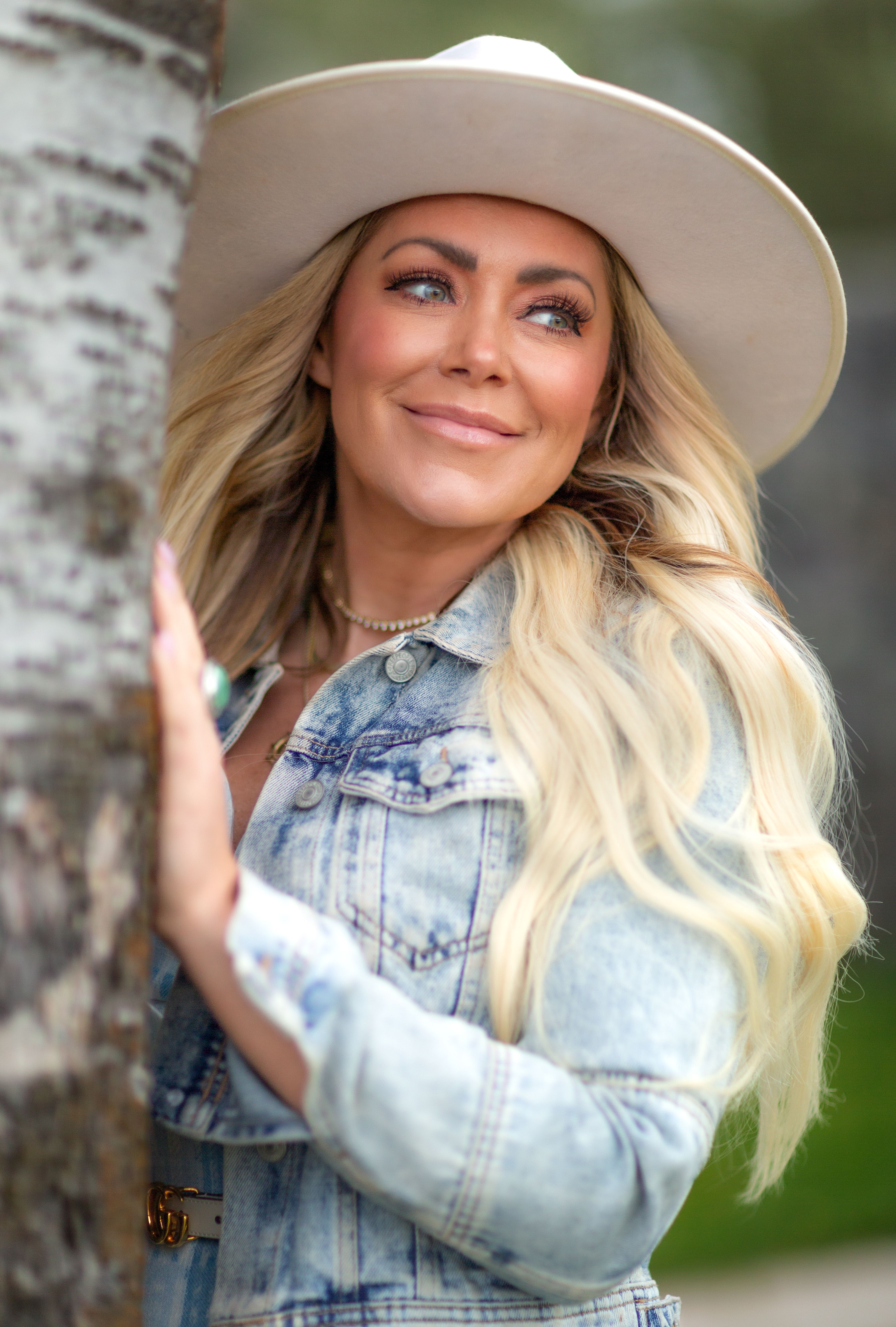 Portrait of a woman with long blonde hair and hat posing by a tree.