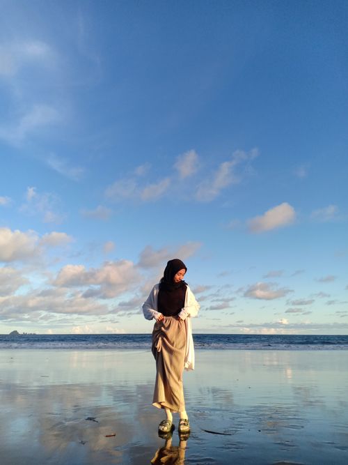Free A woman in a hijab stands on a serene beach under a bright blue sky. Stock Photo