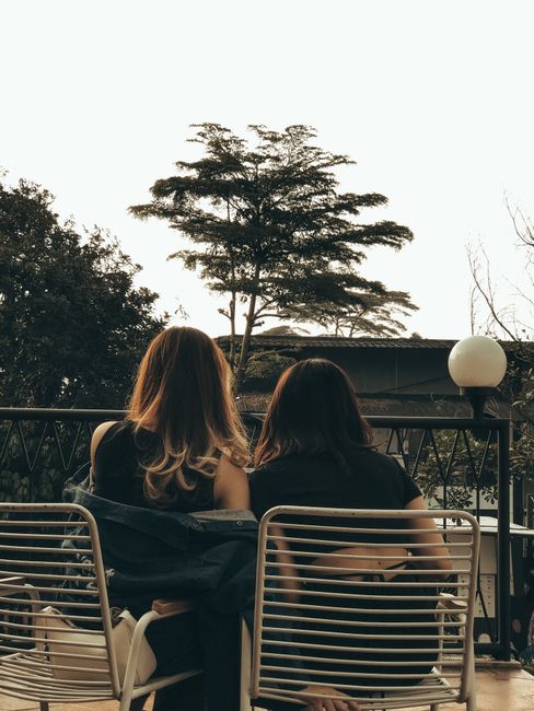 Two women sit on a balcony, sharing a conversation amidst nature.