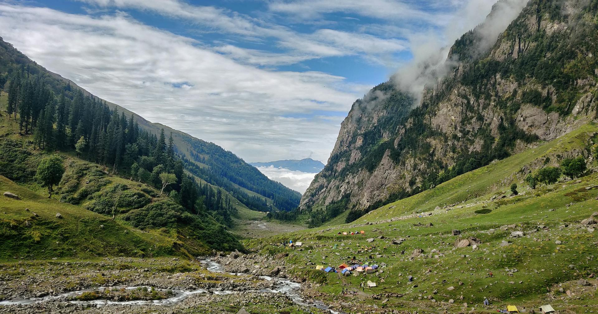 Scenic view of a peaceful camping site in a lush Himalayan valley with tents and a flowing river.