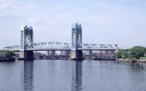 Free A stunning steel bridge across a calm river, framed by a city skyline under a clear blue sky. Stock Photo