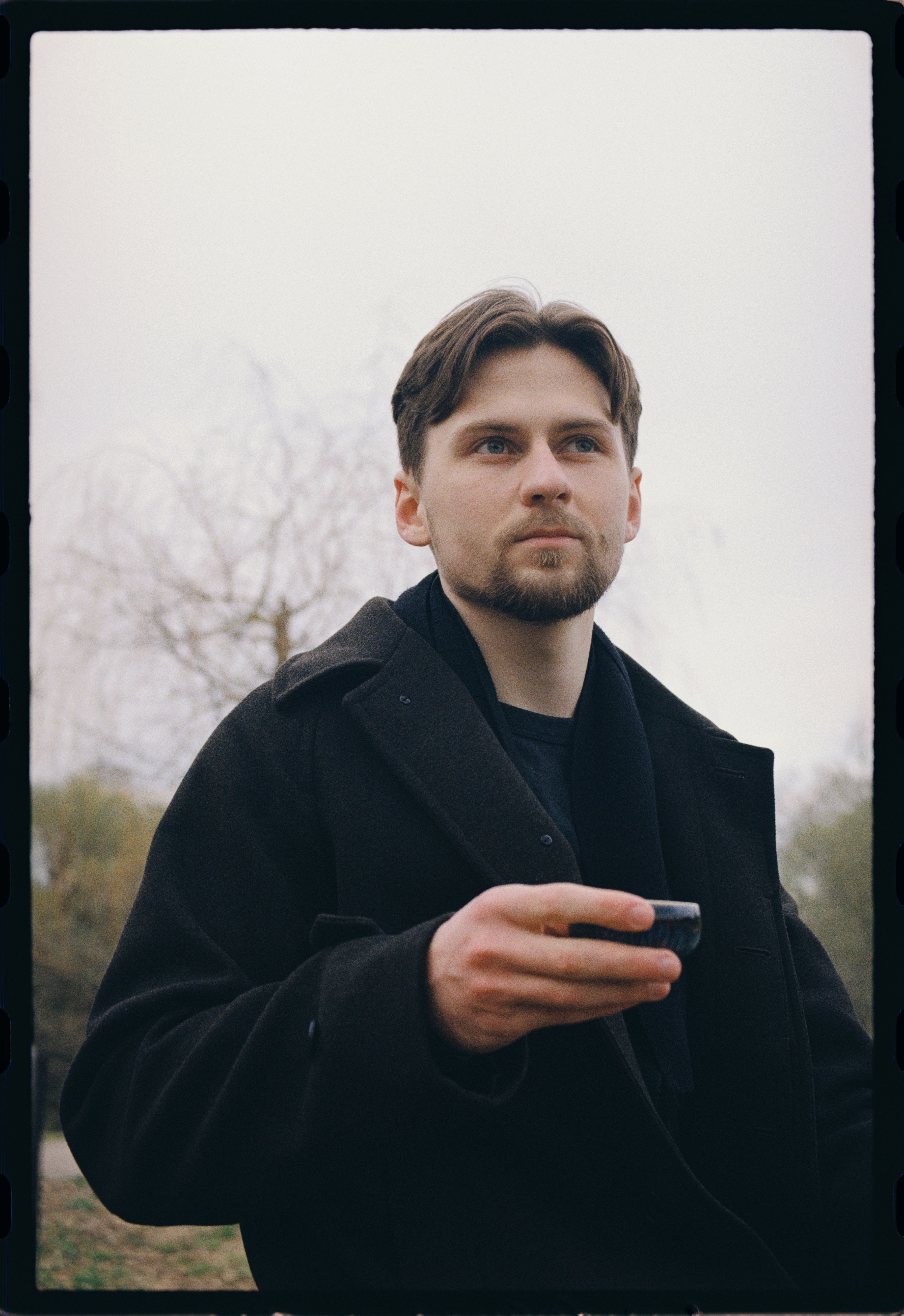 Adult man in a black coat holds a cup outdoors in a Moscow park, showcasing a thoughtful pose.