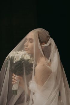 A serene bride under a veil holding a bouquet against a dark backdrop, eyes closed in thought.
