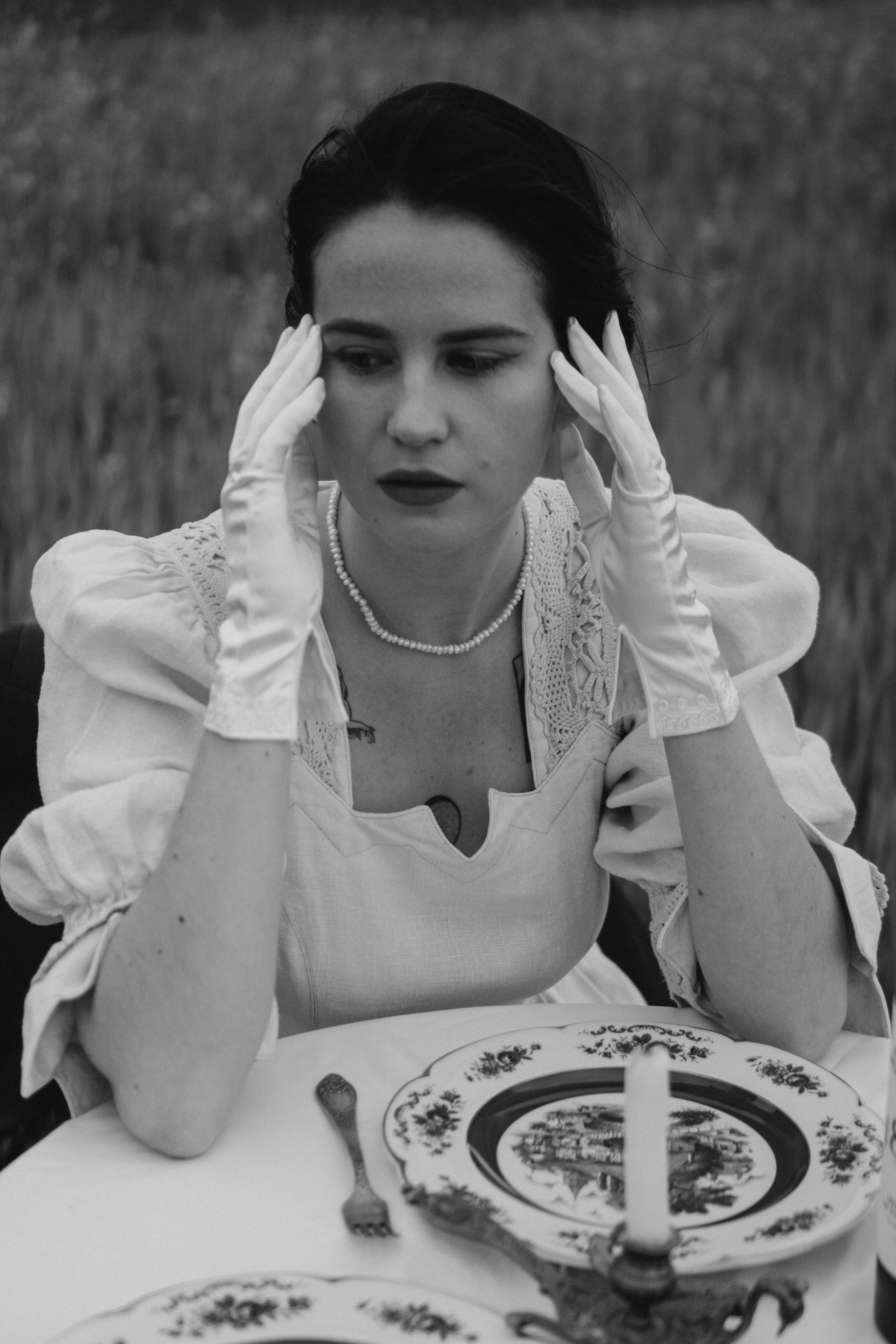 A thoughtful woman in vintage attire sits at a table, captured in black and white.
