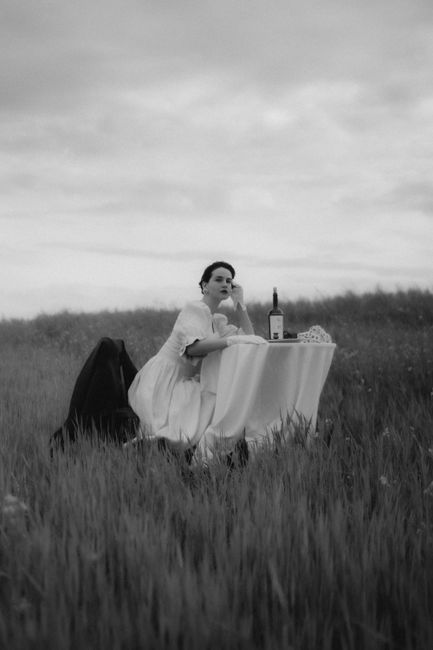 Black and white image of a woman sitting at a table in a meadow, featuring wine.